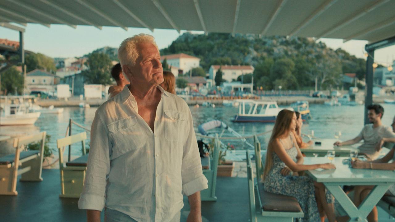 Ivo Batic steht allein in einem offenen Restaurant am kroatischen Meer, im Hintergrund sitzen Gäste an Tischen mit Blick auf Boote.