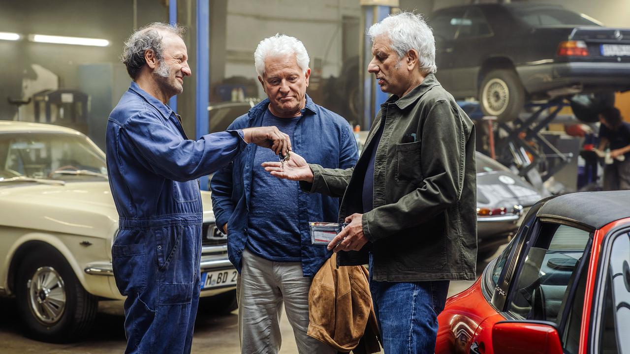 In einer Autowerkstatt zeigt Mechaniker Detlef  zeigt Ivo und Franz ganz stolz den fertigen Porsche und überreicht Franz den Autoschlüssel zwischen abgestellten Fahrzeugen.