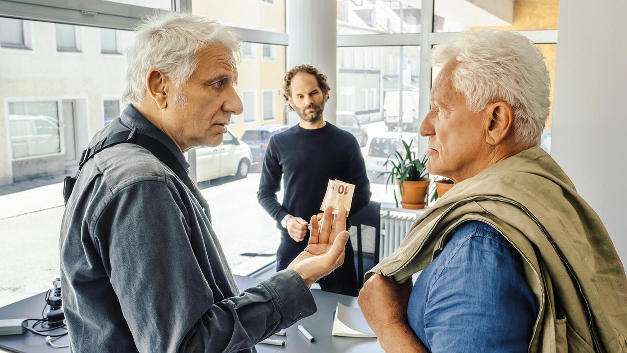 In einem hellen Büro stehen Franz und Ivo vor Stutzki, der für sie einen Personalausweis checken soll. Franz hält einen 10-Euro-Schein in der Hand.