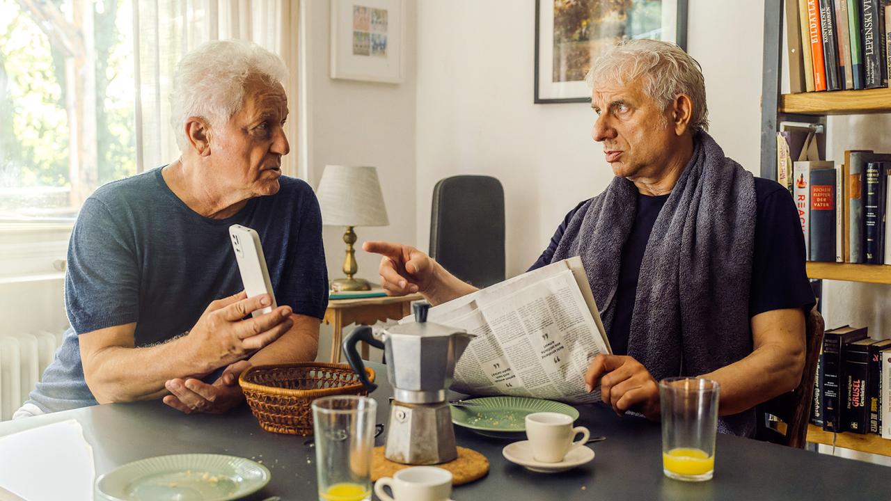 Kriminalhauptkommissar Ivo Batic zeigt Kriminalhauptkommissar Franz Leitmayr beim Frühstück in einer Wohnung das Foto von Zierlinger am Smartphone.
