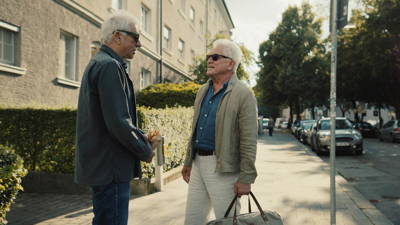 Freudiges Wiedersehen von Franz Leitmayr und Ivo Batic: Sie unterhalten sich auf einem Gehsteig in einem Wohnviertel, Ivo hält dabei eine Tasche in der Hand.