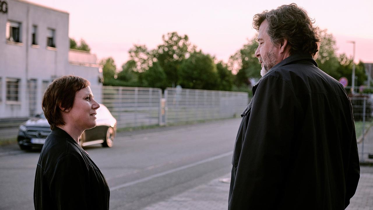 Franziska Tobler und Friedemann Berg stehen sich draußen auf einer ruhigen Straße bei Abendlicht gegenüber und blicken sich an.