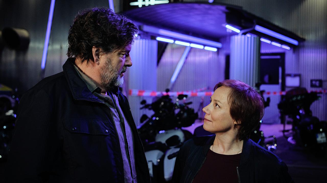 Friedemann Berg  und Franziska Tobler stehen nachts vor einem abgesperrten Gebäude mit Motorrädern und blauem Neonlicht.