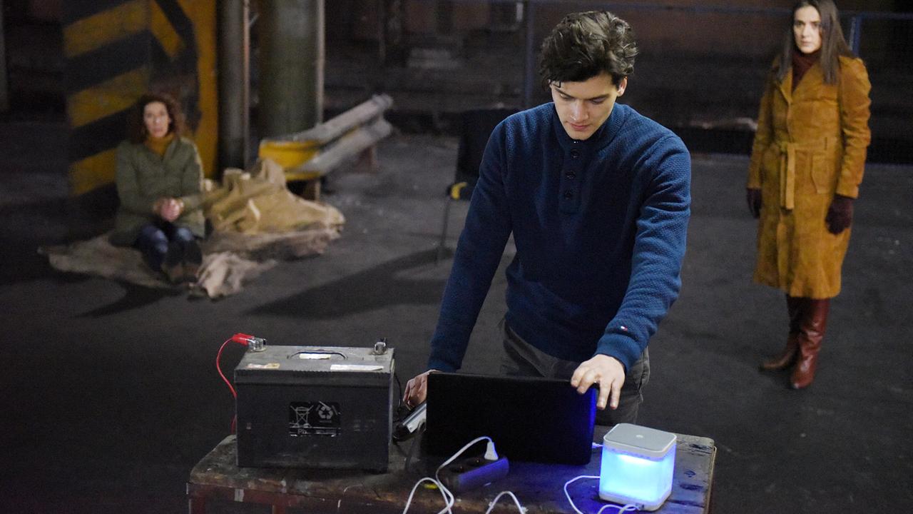 In einer verlassenen Halle bedient David Frank technische Geräte auf einem Tisch, während Bibi Fellner im Hintergrund am Boden sitzt und an einer Wand lehnt und Sarah Adler abseits steht.  
