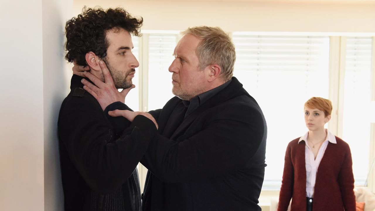 Moritz Eisner drückt Kerem Sami Shafka in einer hellen Wohnung gegen die Wand, während Claudia Eisner im Hintergrund die Szene beobachtet.