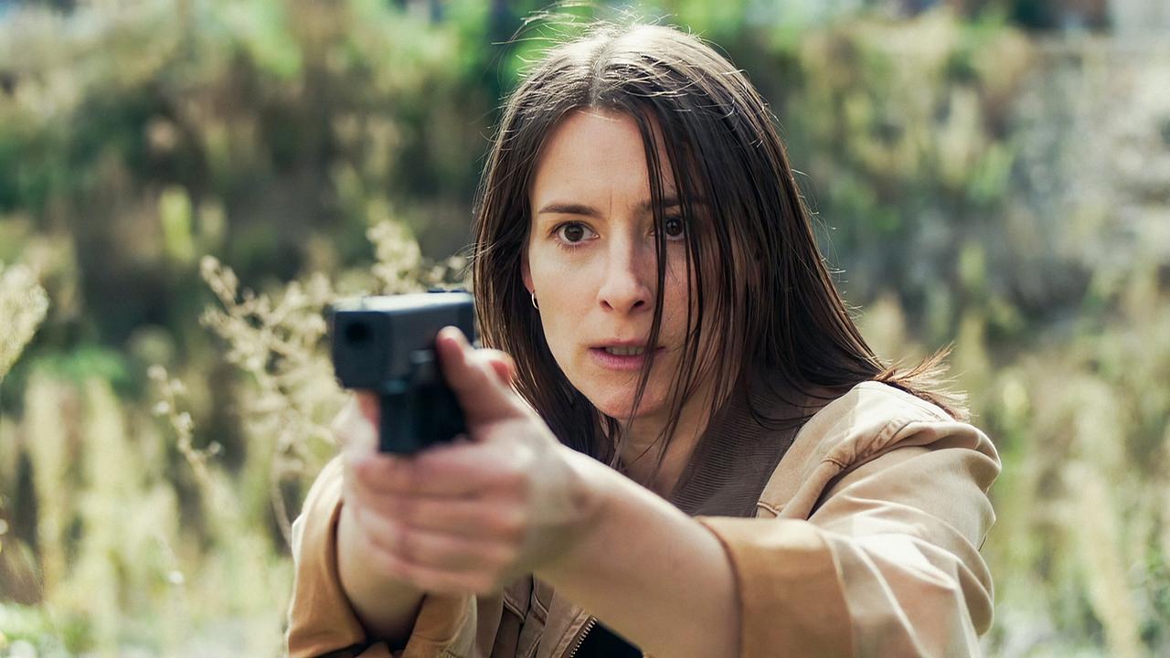 Meret Schande richtet im Freien eine Pistole mit ausgestreckten Armen nach vorne, umgeben von unscharfer Vegetation.