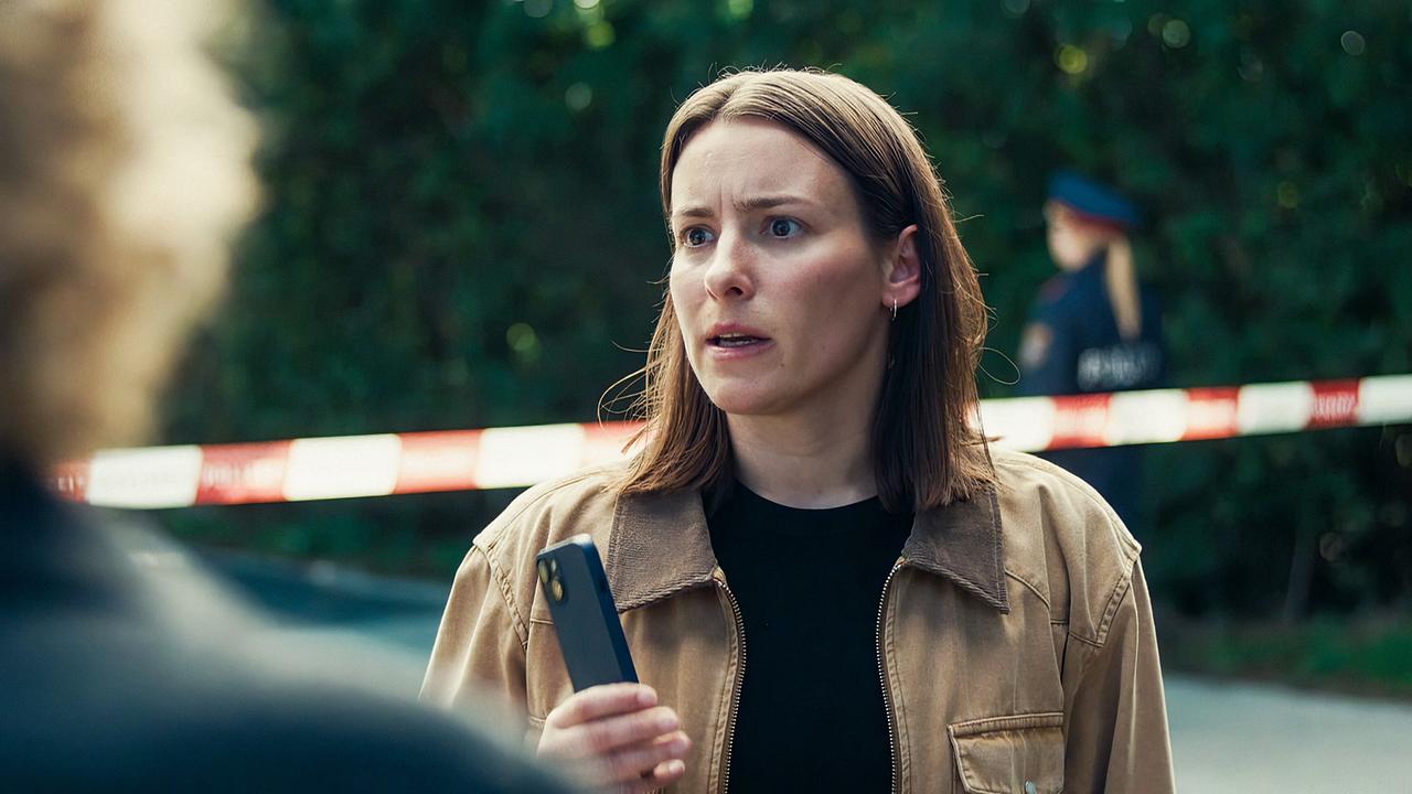 Meret Schande steht mit einem Smartphone in der Hand vor einem Polizeiabsperrband im Freien vor Bibi Fellner, die unscharf im Vordergrund zu sehen ist.
