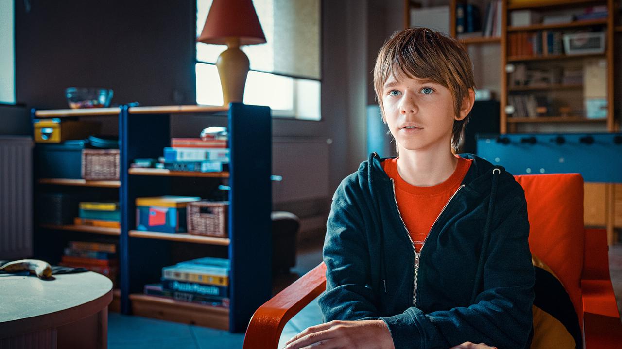 Der junge Levi Hubner sitzt in einem Wohnzimmer auf einem Sessel, mit Regalen und Möbeln im Hintergrund.