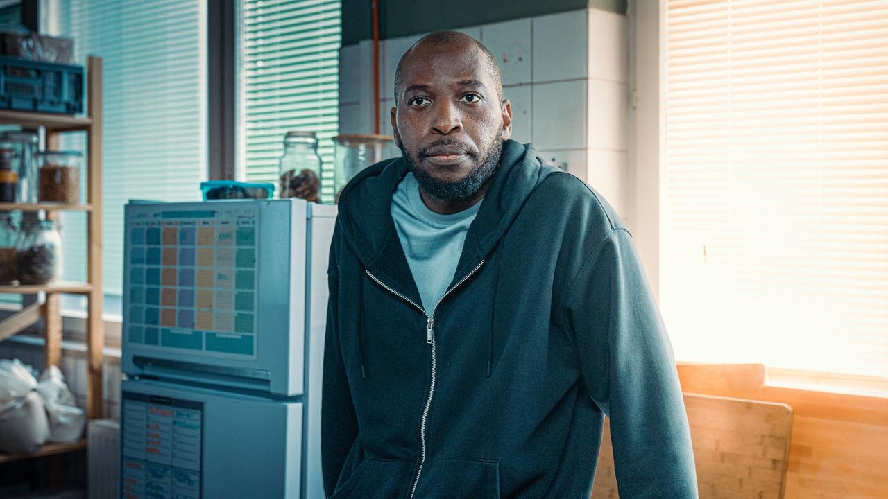 Femi Olaifa lehnt in einer Großküche an einer Arbeitsfläche, umgeben von Geräten und Regalen.