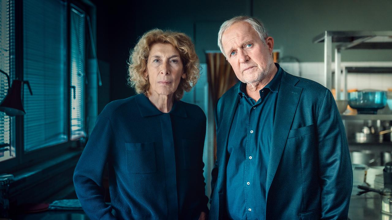 Bibi Fellner und Moritz Eisner stehen nebeneinander in einer abgedunkelten Küche und blicken nach vorne.