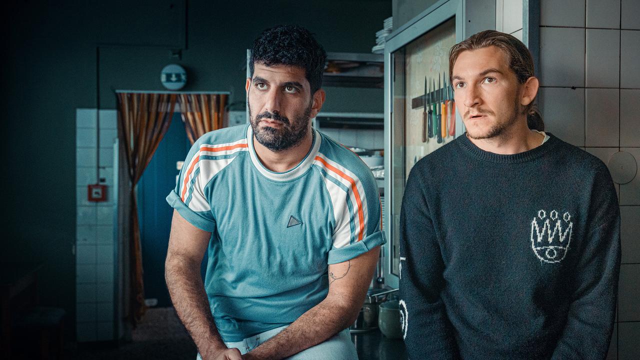 Die Betreuer Araz Barzani und Simon Rechnik lehnen in einer Küche auf einer Arbeitsfläche, im Hintergrund sind Messer und Küchengeräte zu sehen.
