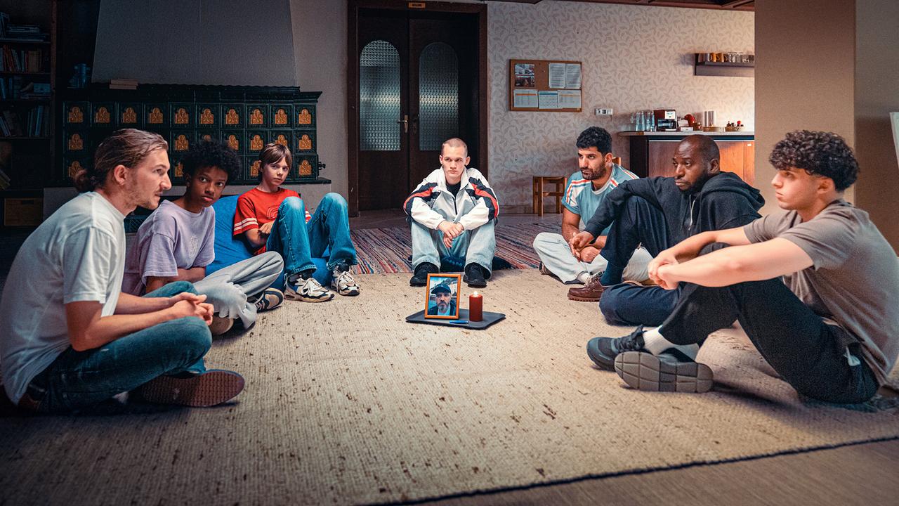 Simon Rechnik, Oki Okotho Sialo, Levi Hubner, Leon Prammer, Araz Barzani, Femi Olaifa und Mo Mohammed Essa sitzen im Kreis auf dem Boden eines Wohnraums, in der Mitte steht ein Tablett mit einer Kerze und einem gerahmten Foto.