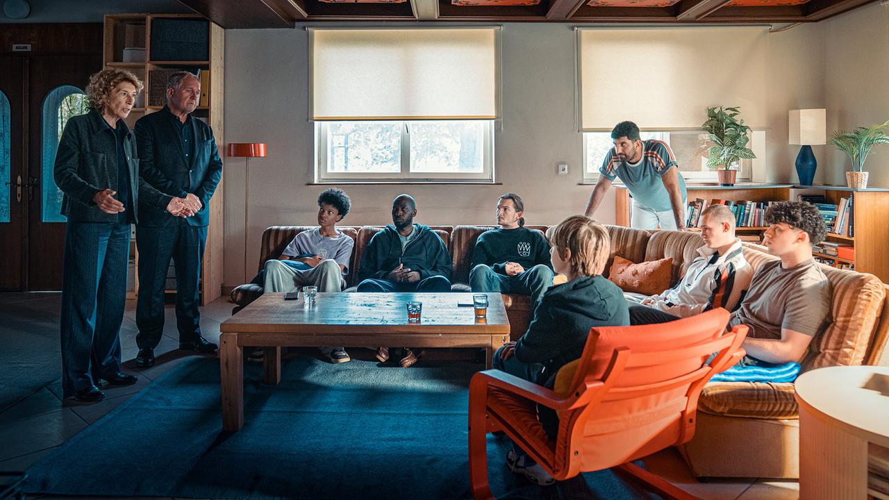 Bibi Fellner und Moritz Eisner stehen im Gemeinschaftsraum des Sonnenhofs vor den auf Sofas an einem Coachtisch sitzenden jugendlichen Bewohnern. Betreuer Femi Olaifa steht dabei. 