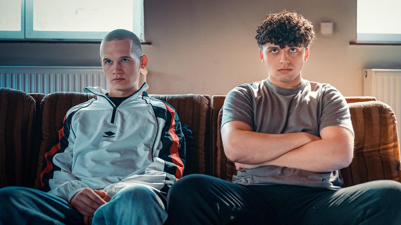 Die Bewohner Leon Prammer und Mo Mohammed Essa mit verschränkten Armen sitzen nebeneinander auf einem Sofa im Gemeinschaftsraum.