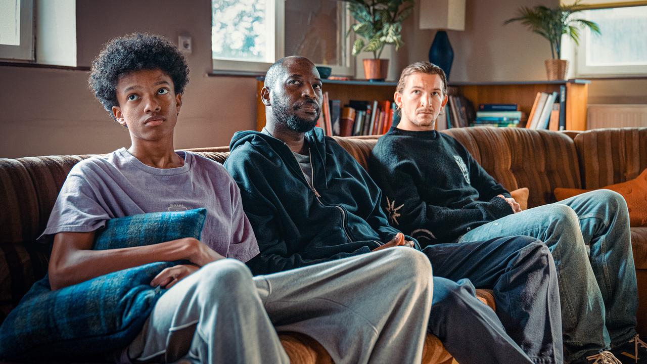 Oki Okotho Sialo, Femi Olaifa und Simon Rechnik  sitzen nebeneinander auf einem Sofa in einem Gemeinschaftsraum mit Pflanzen und Bücherregalen im Hintergrund.