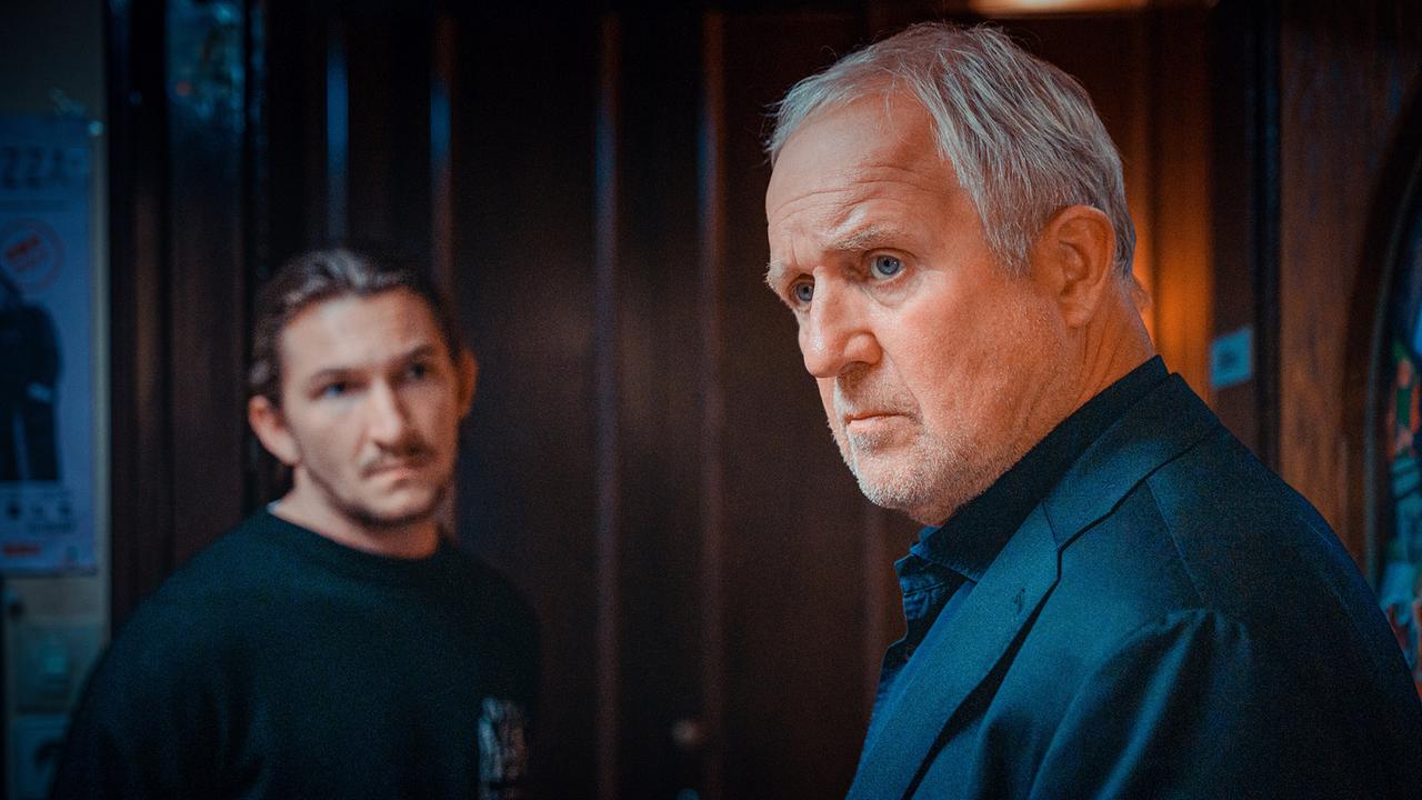 Moritz Eisner steht neben Simon Rechnik in einem Flur nahe einer dunklen Holztür.