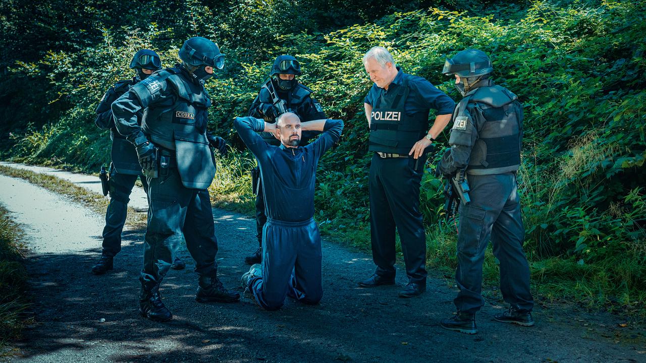  Mehrere bewaffnete Einsatzkräfte unter ihnen Moritz Eisner stehen um den knienden Jürgen Siller auf einem Waldweg.