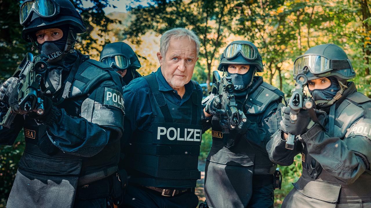  Bewaffnete Einsatzkräfte mit Moritz Eisner in Schutzkleidung in der Mitte stehen in einem bewaldeten Gebiet.