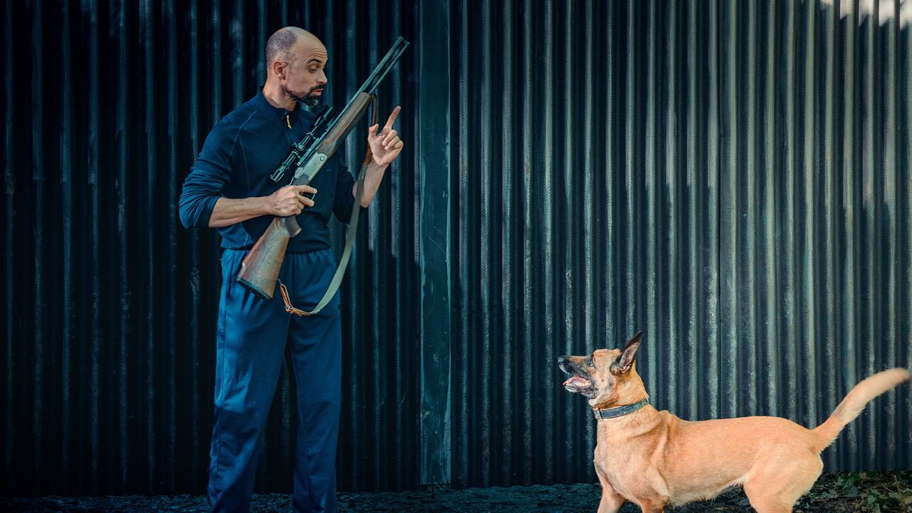 Jürgen Siller steht mit einem Gewehr in der Hand vor einer Wellblechwand und gibt einem Hund vor ihm ein Handzeichen.