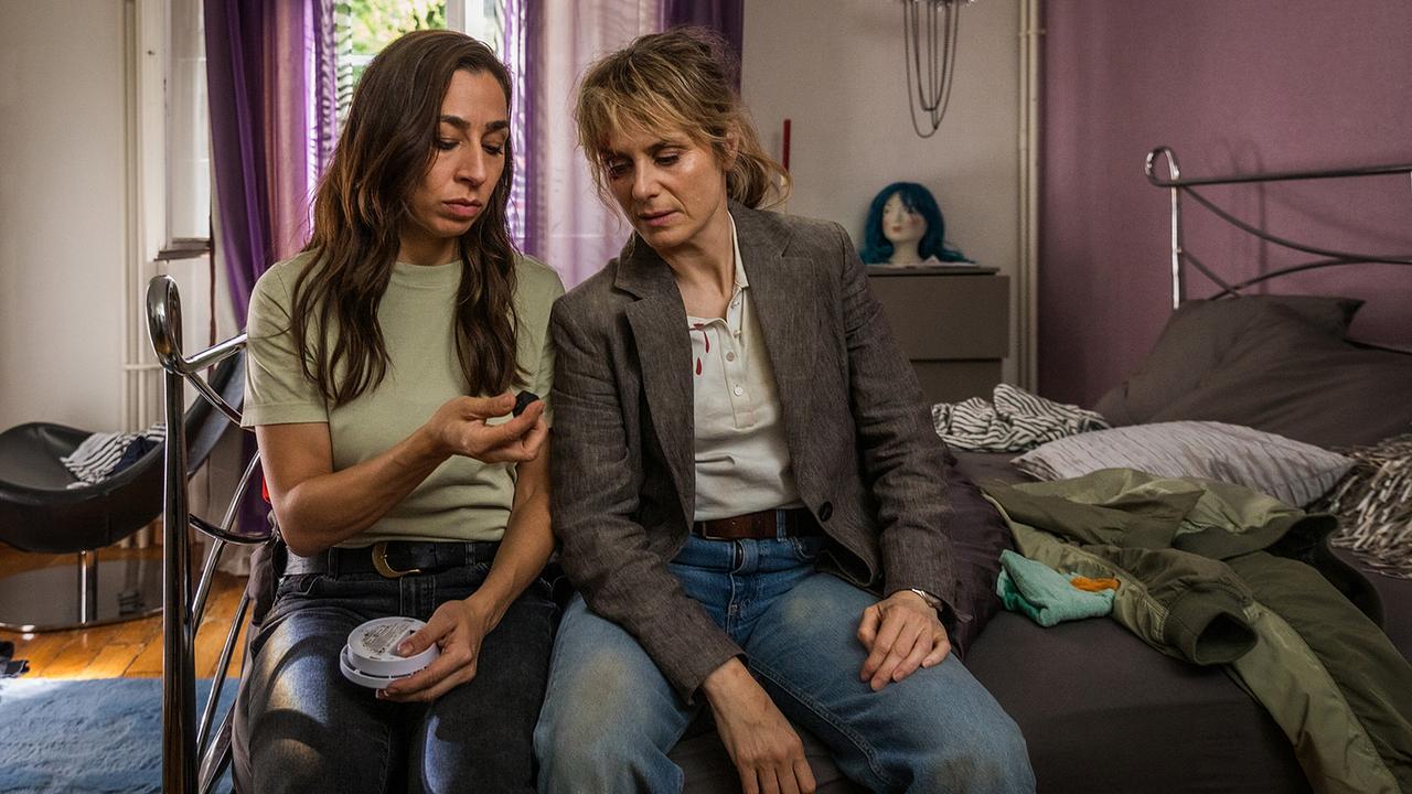 Kommissarin Tessa Ott und ihre Kollegin Kommissarin Isabelle Grandjean sitzen am Bett in der Wohnung des Opfers und betrachten einen kleinen Gegenstand.