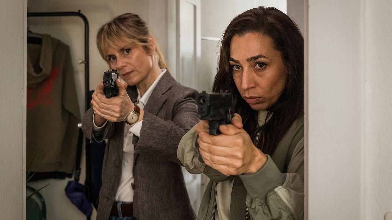 Bekommen überraschenden Besuch in der Wohnung des Opfers: Kommissarin Tessa Ott und Kommissarin Isabelle Grandjean zielen mit gezogenen Pistolen aus einem engen Raum heraus.