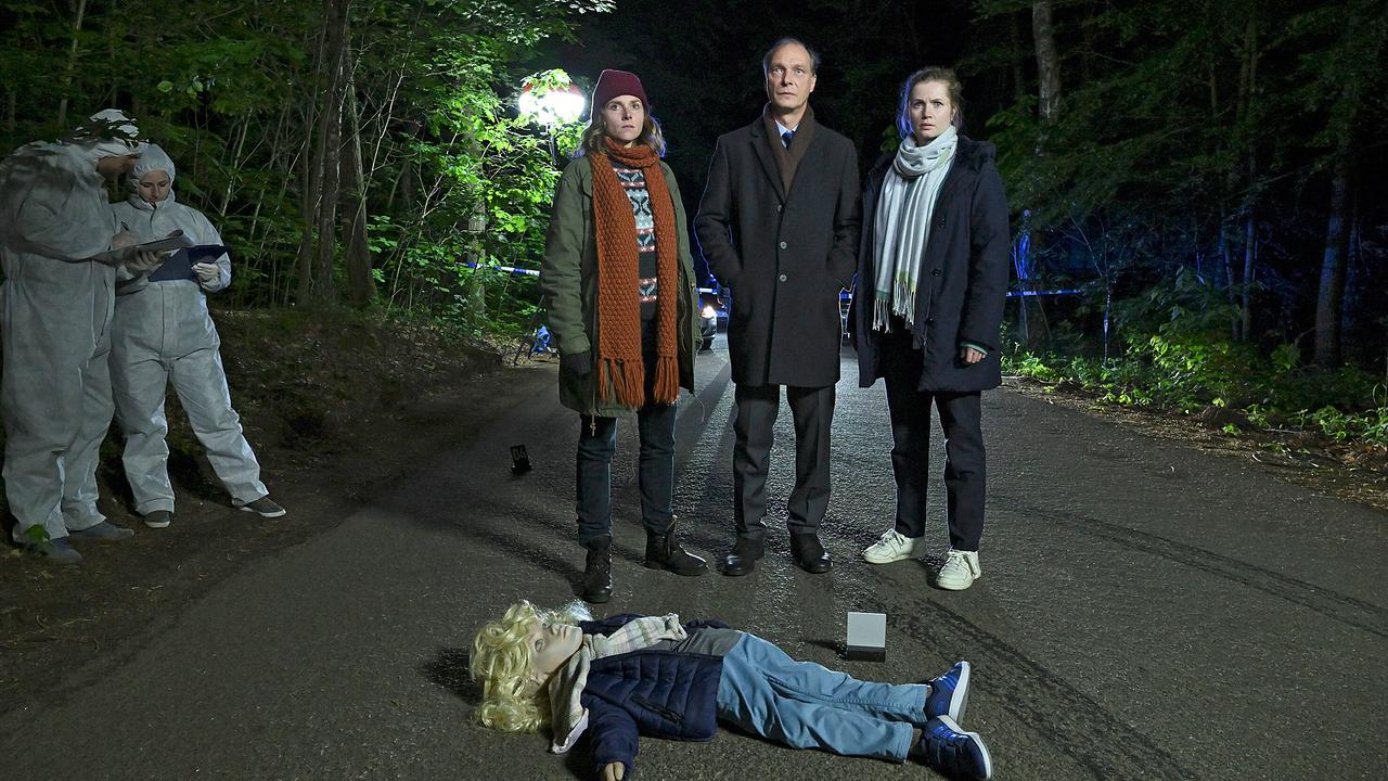 Das Ermittlerteam Karin Gorniak, Peter Michael Schnabel und Leo Winkler steht nachts auf einer Landstraße vor einem abgesperrten Bereich, während eine Attrappe in der Form eines Kindes reglos auf der Straße liegt.