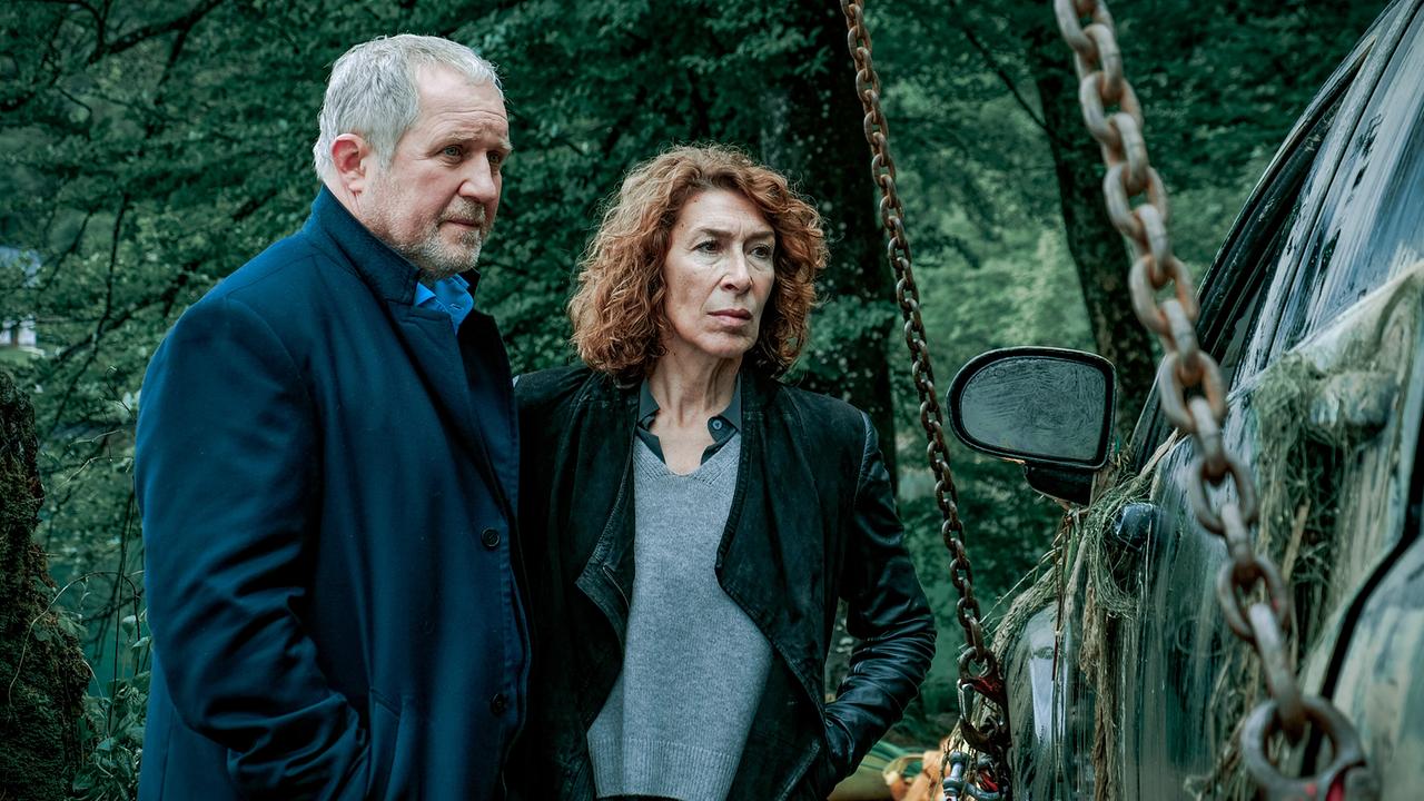 Moritz Eisner und Bibi Fellner betrachten im Wald ein verschmutztes Auto, das an Ketten hängt.