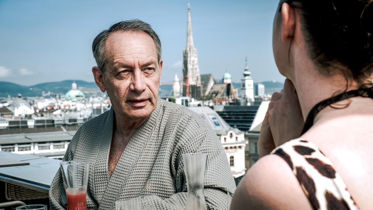Ein älterer Mann David Weimann im Bademantel spricht auf einer Terrasse mit einer jungen Frau Sybille Wildering auf einer Dachterrasse bei Drinks vor der Wiener Skyline.