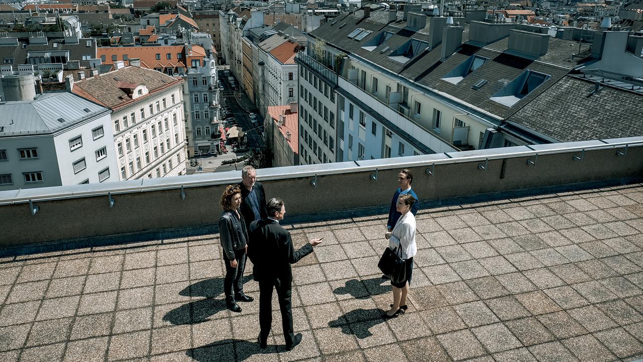 Auf einer großen Dachterrasse stehen Bibi Fellner, Moritz Eisner und Oberst Ernst Rauter gegenüber von Dr. Maria Digruber und Lukas Kragl und unterhalten sich.