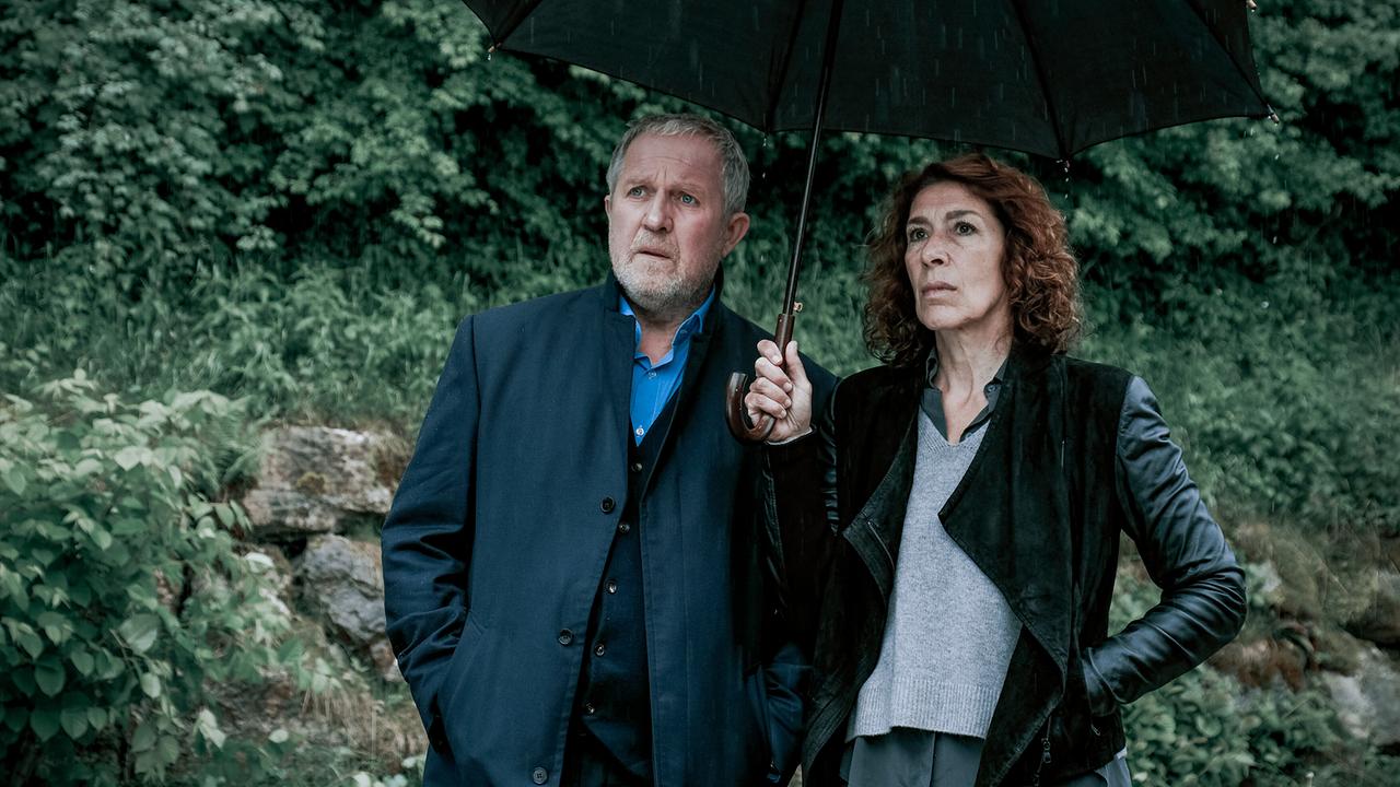 Moritz Eisner und Bibi Fellner stehen unter einem Regenschirm im nassen Wald und blicken ernst nach vorne.