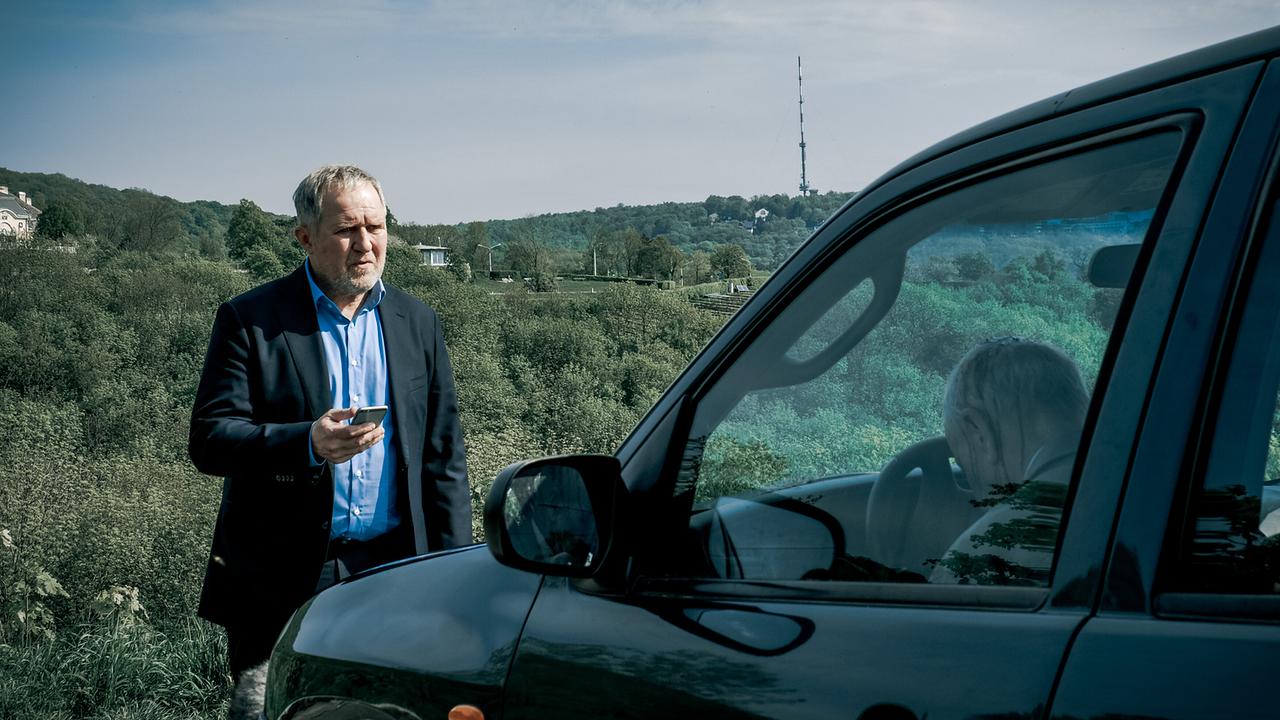 Moritz Eisner steht mit Handy neben einem dunklen Van, in dem jemand reglos mit dem Kopf am Lenkrad sitzt, in ländlicher Umgebung.