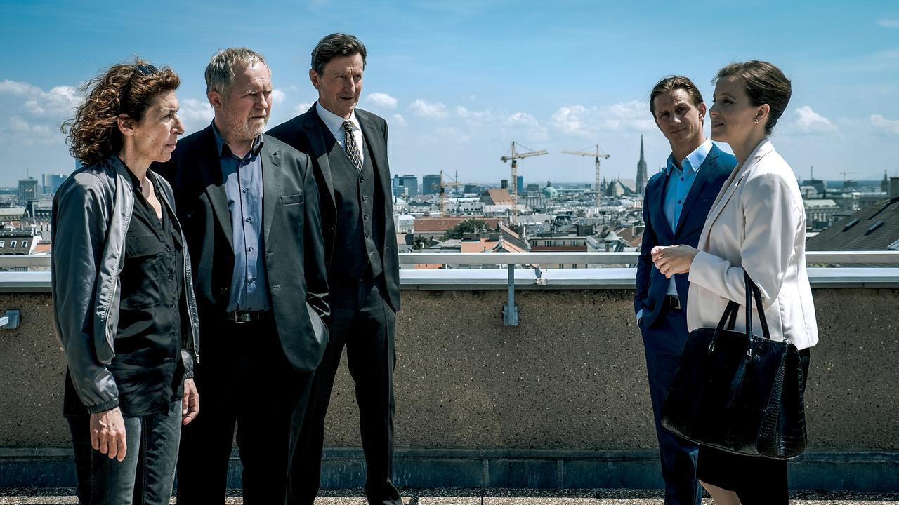 Bibi Fellner, Moritz Eisner und Oberst Ernst Rauter stehen gegenüber von Dr. Maria Digruber und Lukas Kragl auf einer Dachterrasse mit Blick über die Stadt.