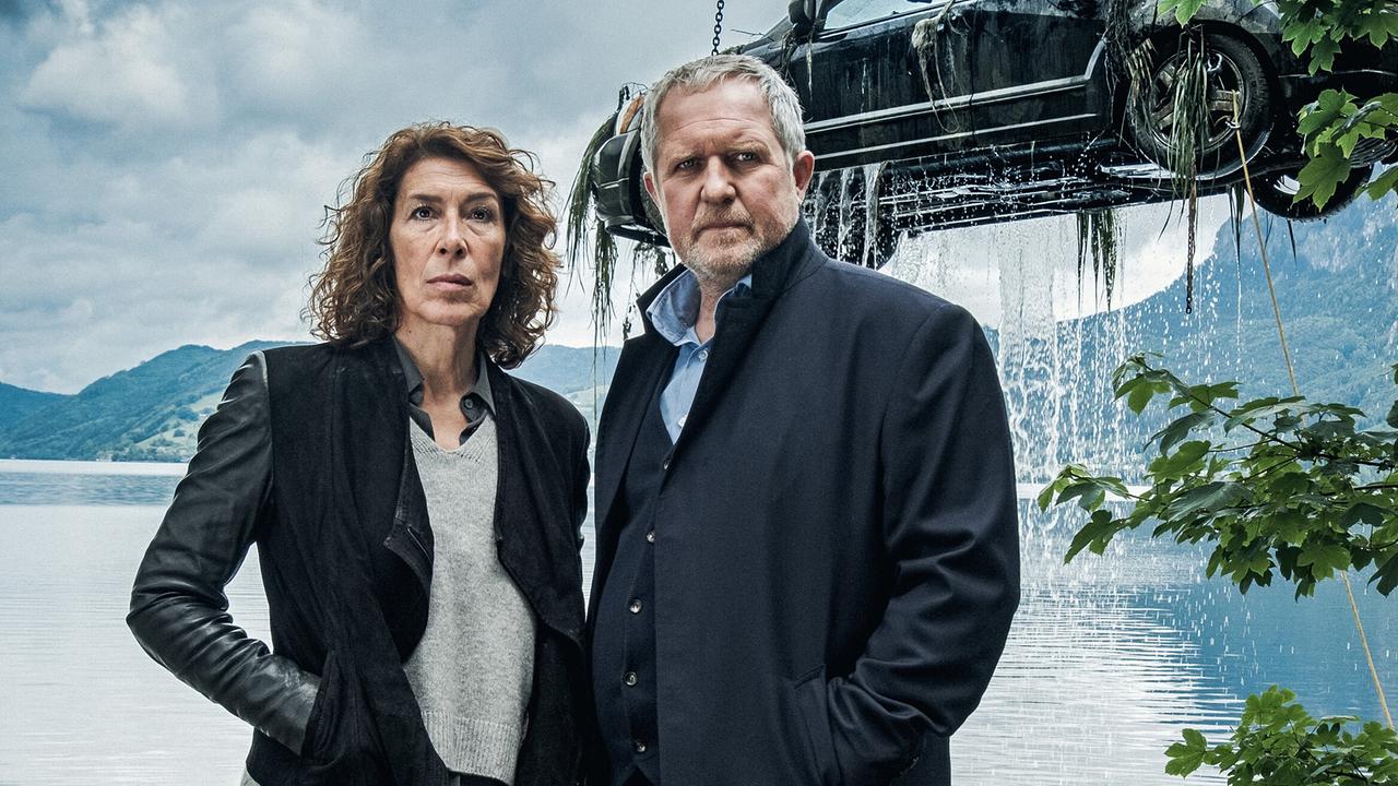 Bibi Fellner und Moritz Eisner vor einem See, während im Hintergrund ein Auto aus dem Wasser gehoben wird.