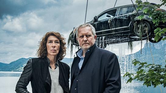 Bibi Fellner und Moritz Eisner vor einem See, während im Hintergrund ein Auto aus dem Wasser gehoben wird.