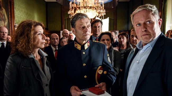 © ORF/Hubert Mican Im prunkvollen Saal mit mehreren Personen steht im Vordergrund Stefan Pohl in Uniform mit goldenen Knöpfen und roten Abzeichen hält eine rote Schmuckbox und einen Umschlag, während Bibi Fellner und Moritz Eisner links und rechts neben ihm stehen.