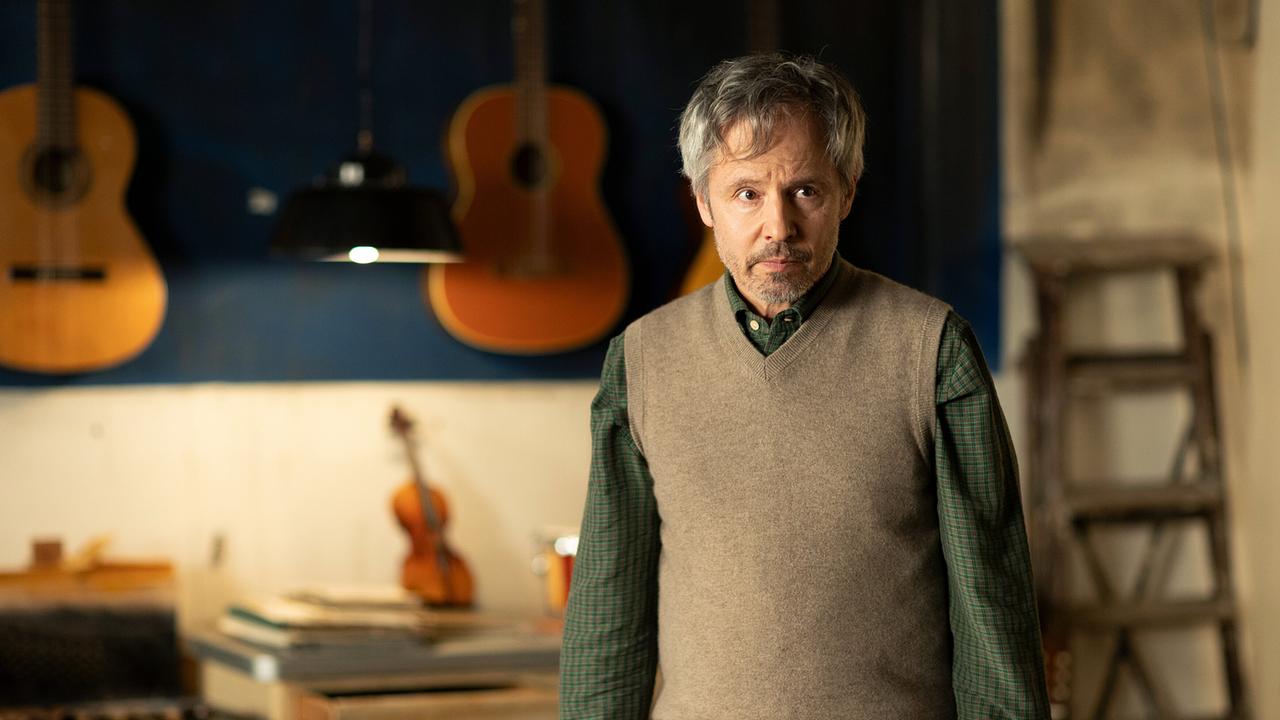 Herr Miller, Leiter der Musikschule „König“, steht ernst vor mehreren an der Wand hängenden Gitarren.
