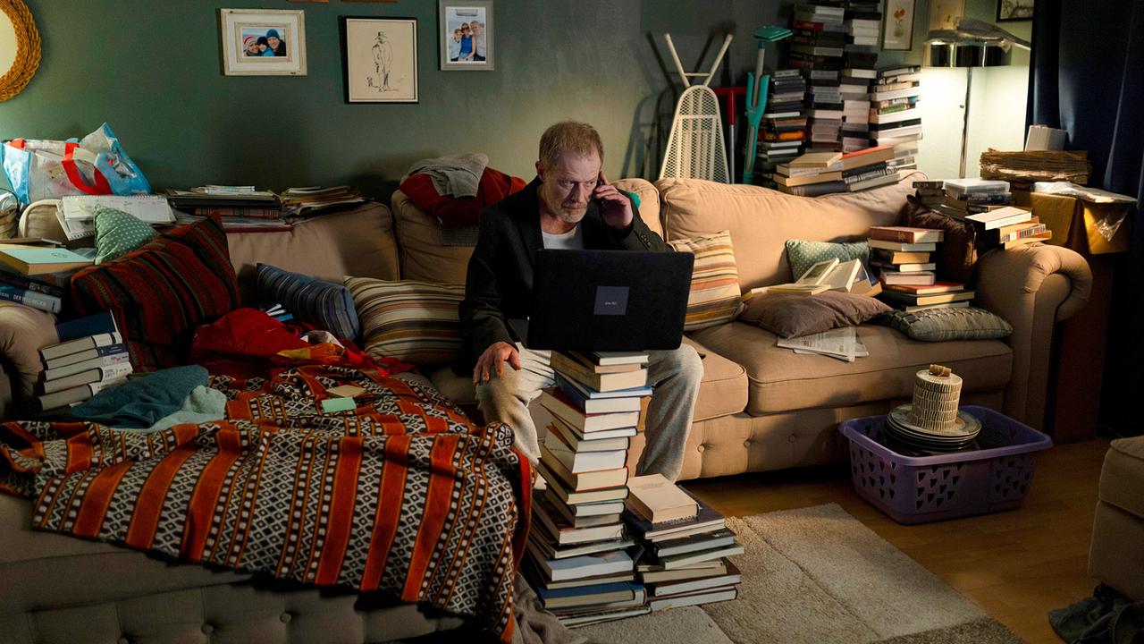 Matthias Vogel sitzt mit einem Laptop in einem überfüllten Wohnzimmer auf dem Sofa zwischen hohen Bücherstapeln.