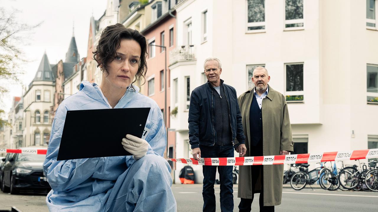 KTU-Chefin Natalie Förster dokumentiert einen abgesperrten Tatort am Rand eines Kölner Stadtparks, während die Kommissare Max Ballauf und Freddy Schenk im Hintergrund stehen.