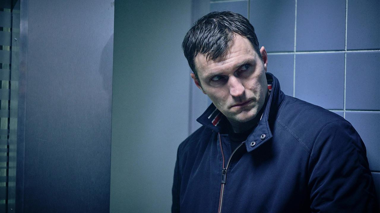 Ulf Meisner mit sichtbar gezeichnetem Gesicht lehnt in einem kühl beleuchteten, gefliesten Raum an einer Wand und  will das Polizeipräsidium erst wieder verlassen, wenn er mit der Kommissarin gesprochen hat.