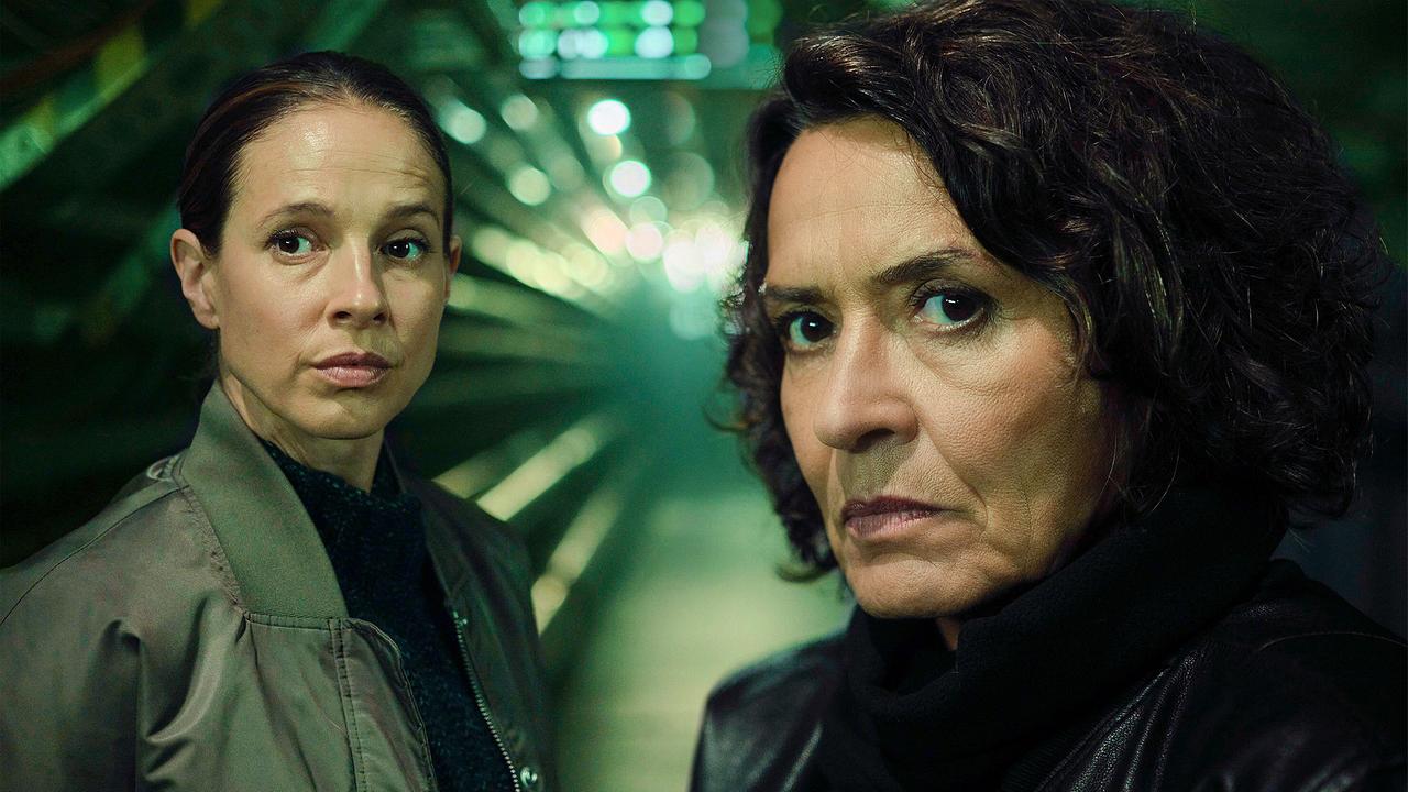 Lena Odenthal und Johanna Stern blicken in einem grünlich beleuchteten, technischen Gang wachsam nach vorne.