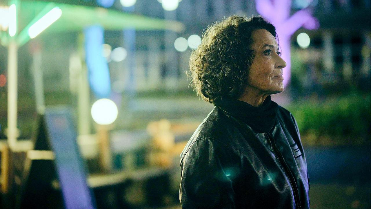 Lena Odenthal in schwarzer Lederjacke steht spöttisch lächelnd in einer neonbeleuchteten Umgebung.