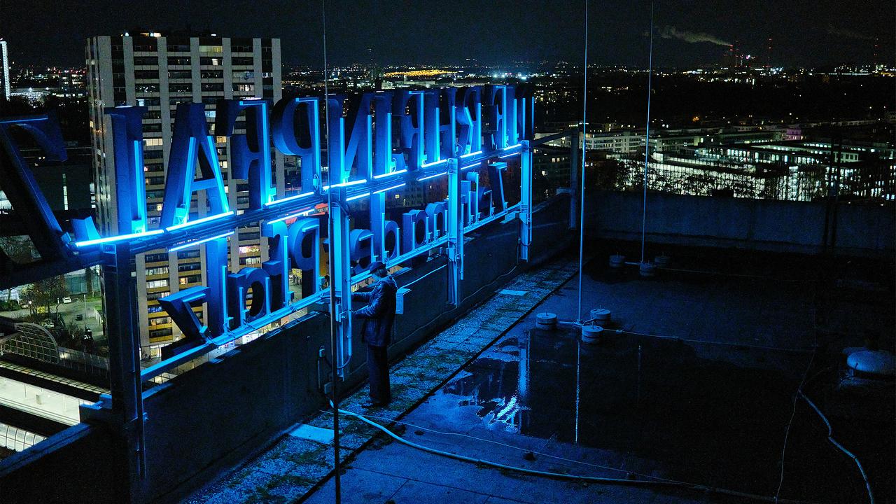 Chris Lohberg holt nachts die Pistole seines Bruders aus einem Versteck auf dem Hochhausdach an einem blau leuchtenden Schriftzug, während im Hintergrund die Stadt zu sehen ist.