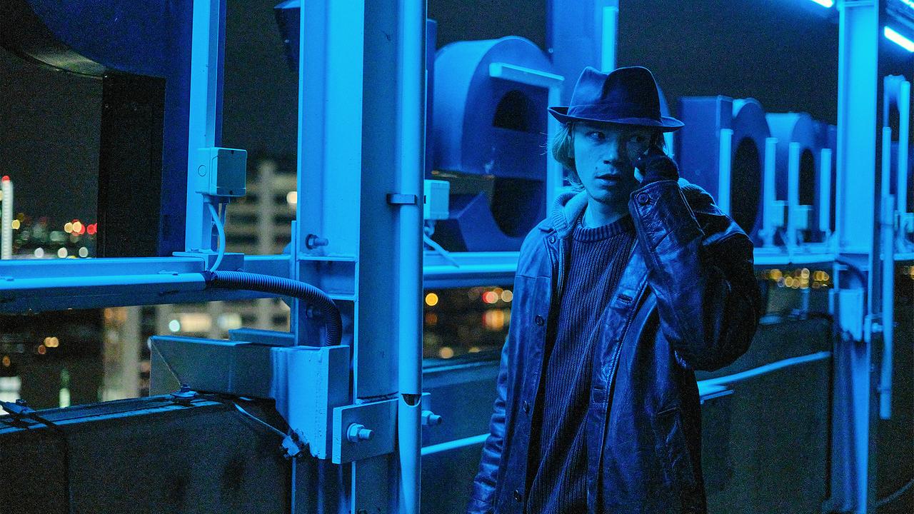Chris mit Hut telefoniert nachts auf einem blau beleuchteten Hochhausdach.