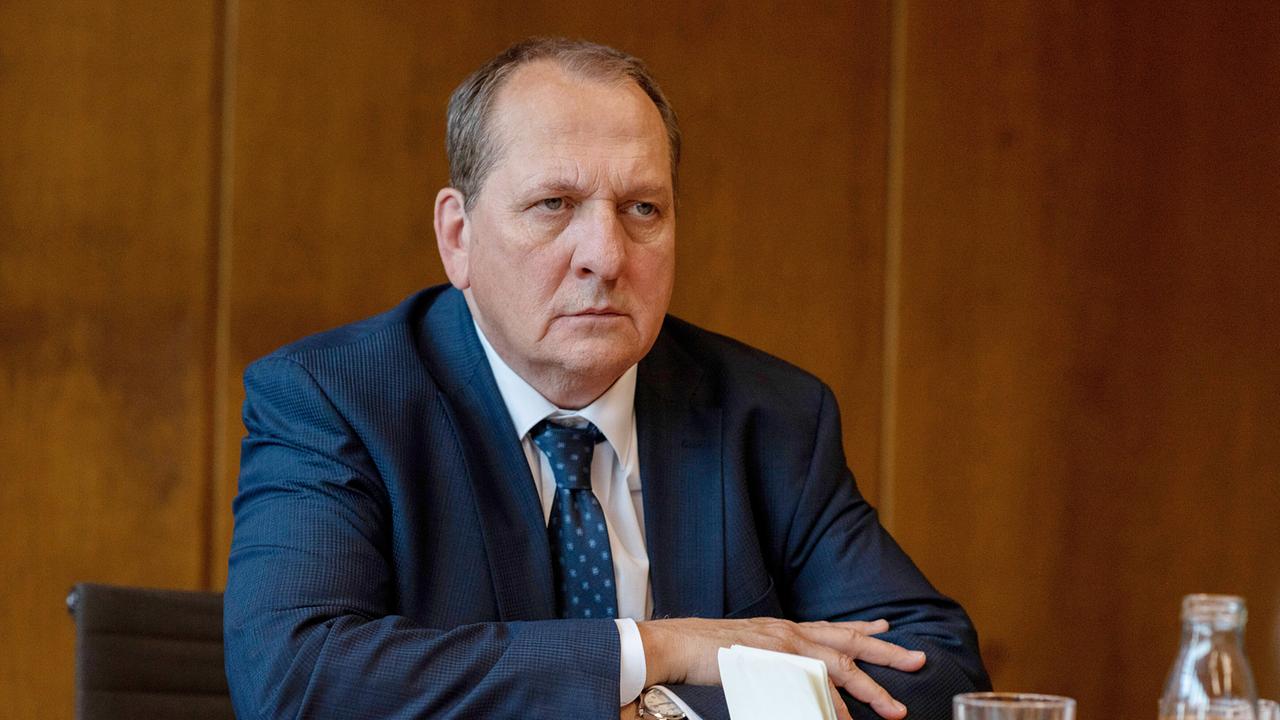 Roland Schladitz im Anzug sitzt mit ernster Miene an einem Tisch in einem holzvertäfelten Raum.