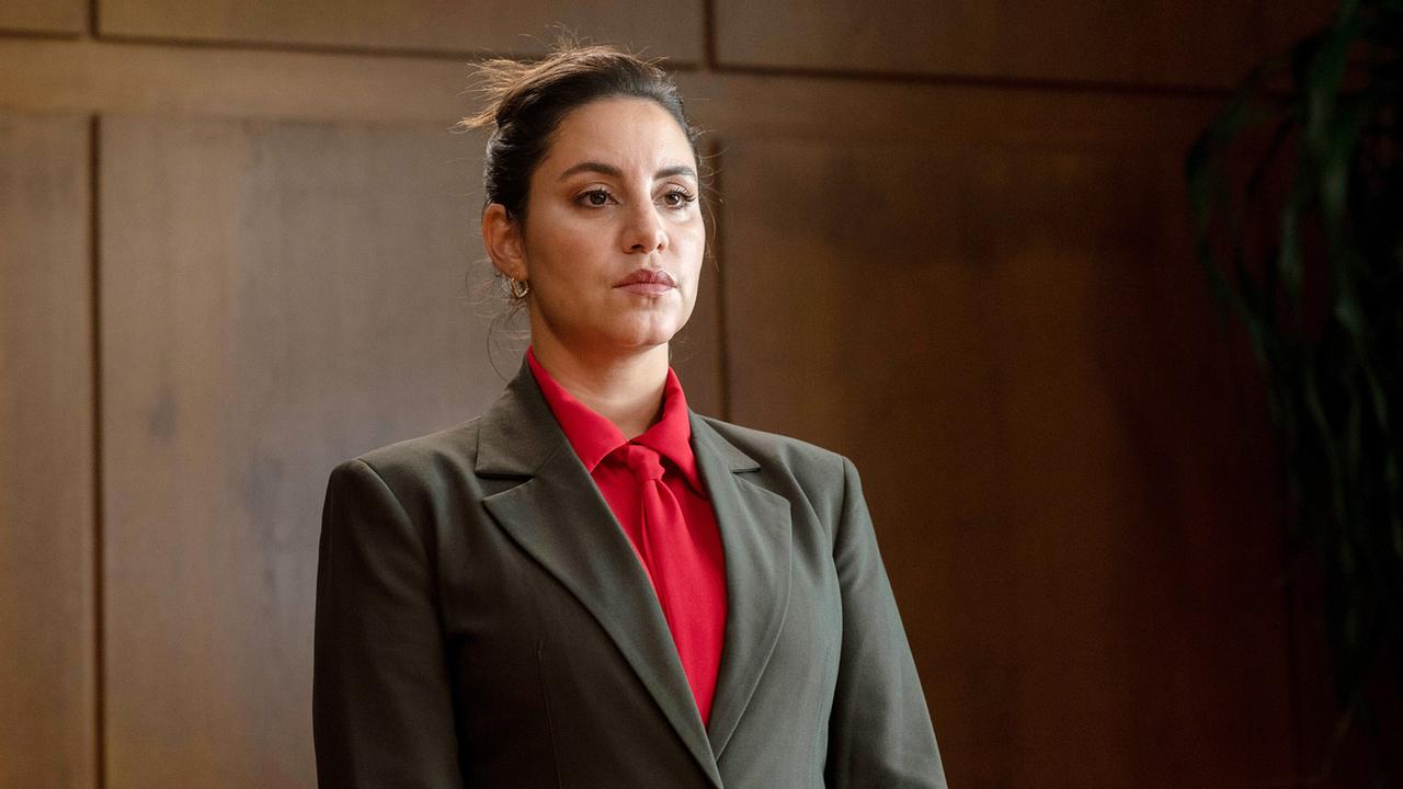 Mila Sahin bewirbt sich als Kriminalrätin und steht mit hochgestecktem Haar, in dunklem Blazer über einer roten Bluse ernst vor einer holzvertäfelten Wand.