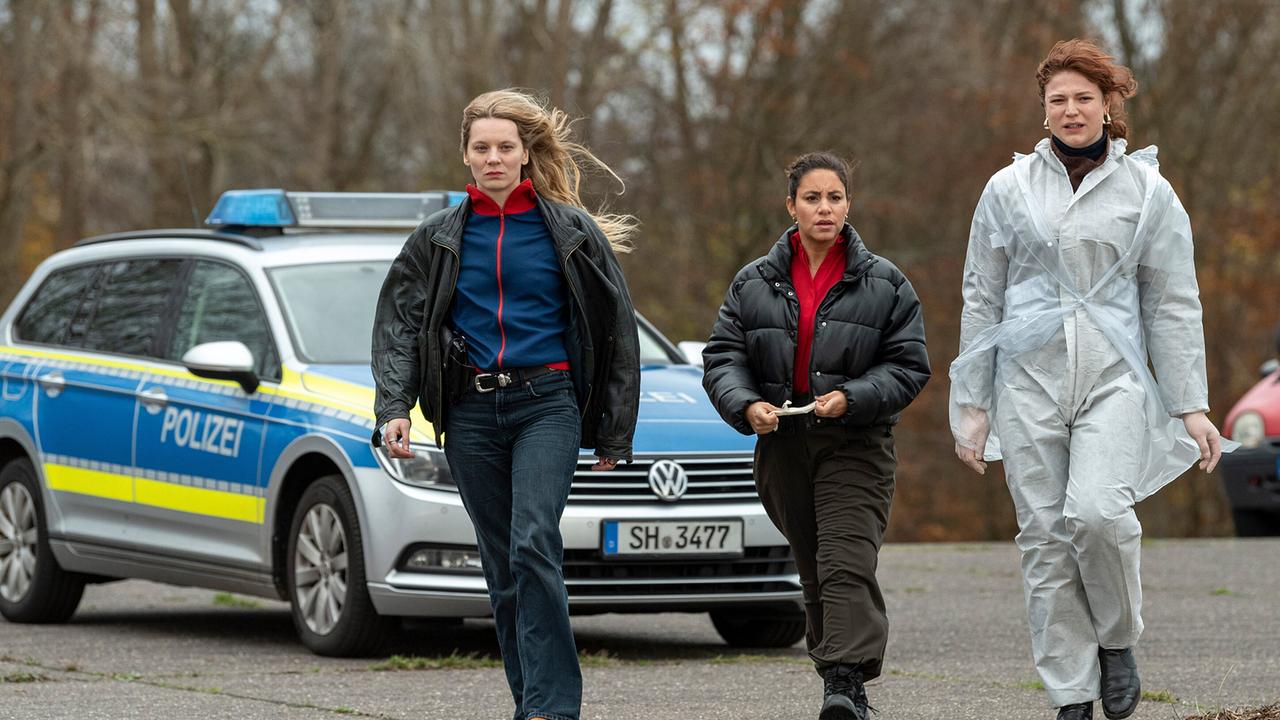 Mila, ihre neue Kollegin Anna Nowak und eine Spurensicherin gehen vor einem Polizeiauto zum Tatort im Freien.