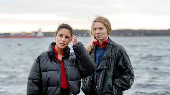Mila und Anna Nowak stehen bei windigem Wetter am Wasser, während im Hintergrund eine Fähre vorbeifährt.