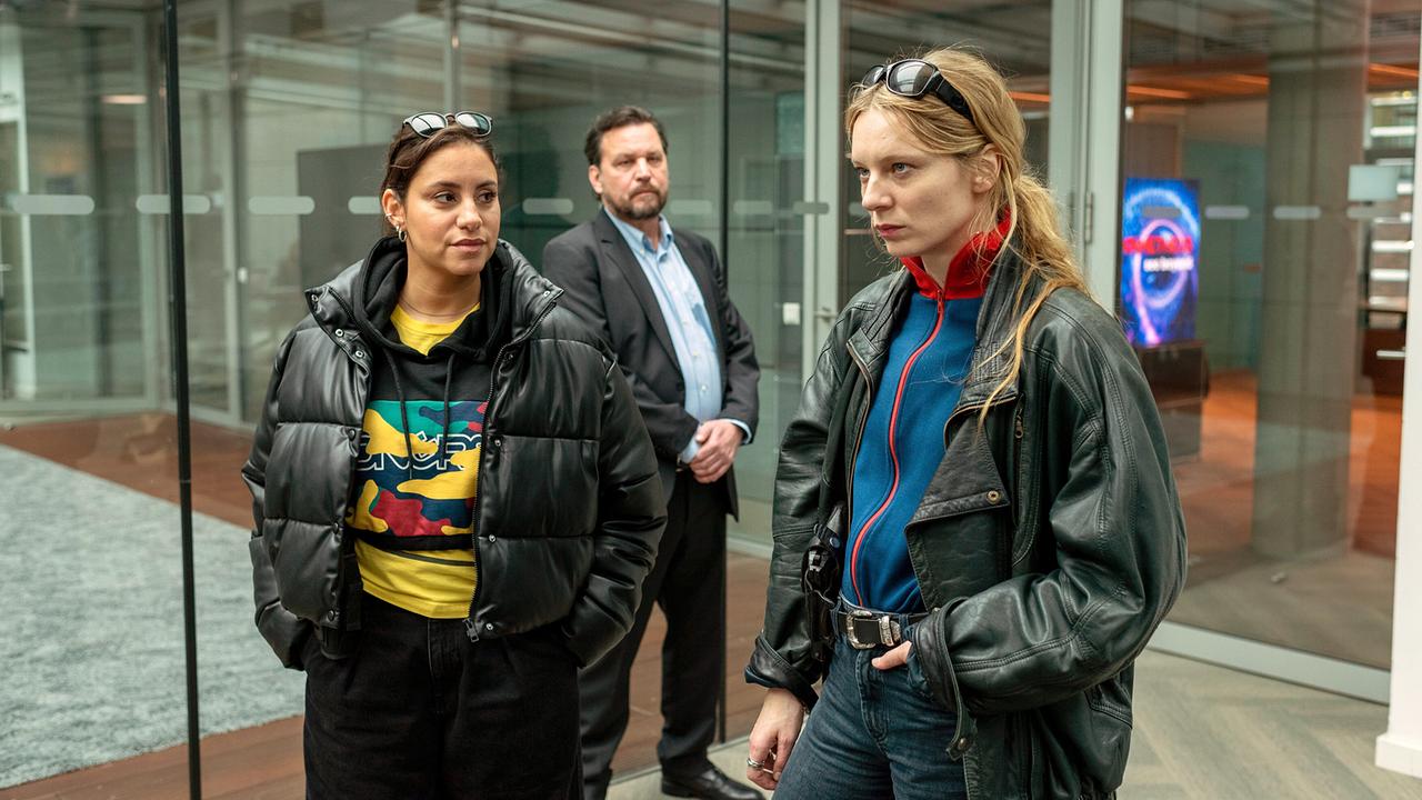 Mila Sahin und Anna Nowak gehen in einem modernen Büro in einem Glasflur an Franco Steiner vorbei.