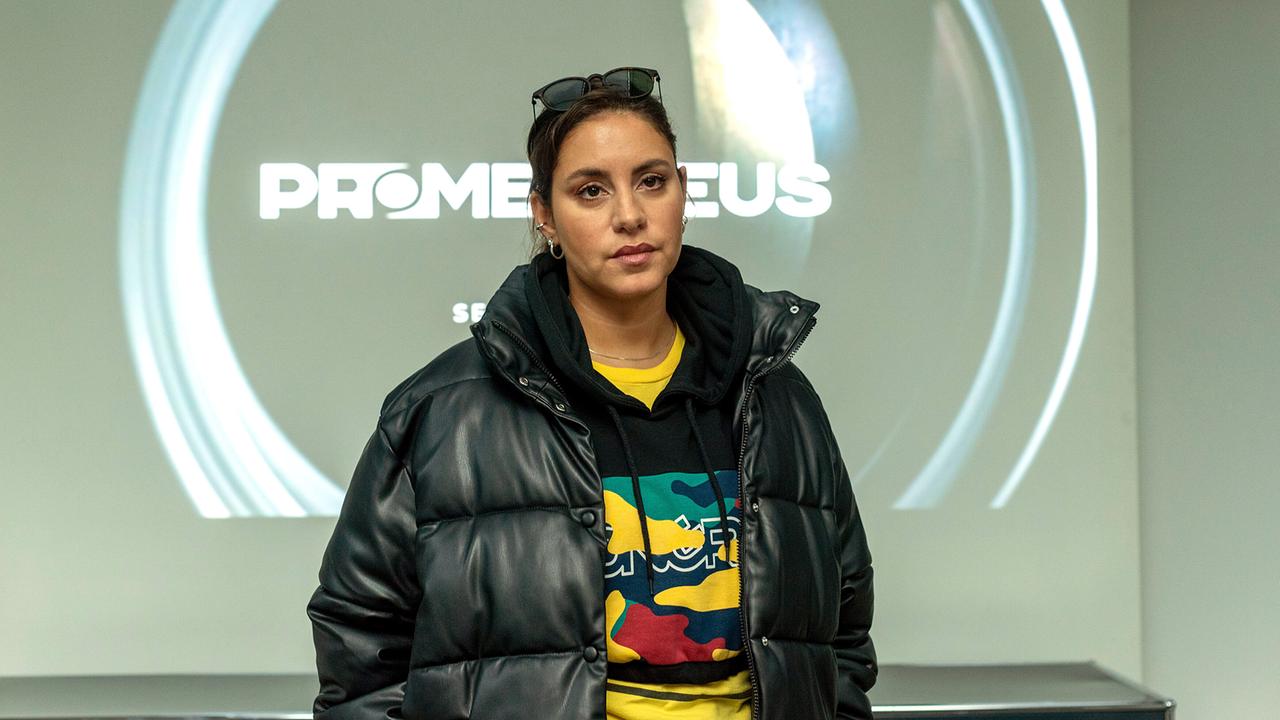 Mila Sahin in schwarzer Steppjacke steht vor einer Wandprojektion mit der Aufschrift „PROMETHEUS“.