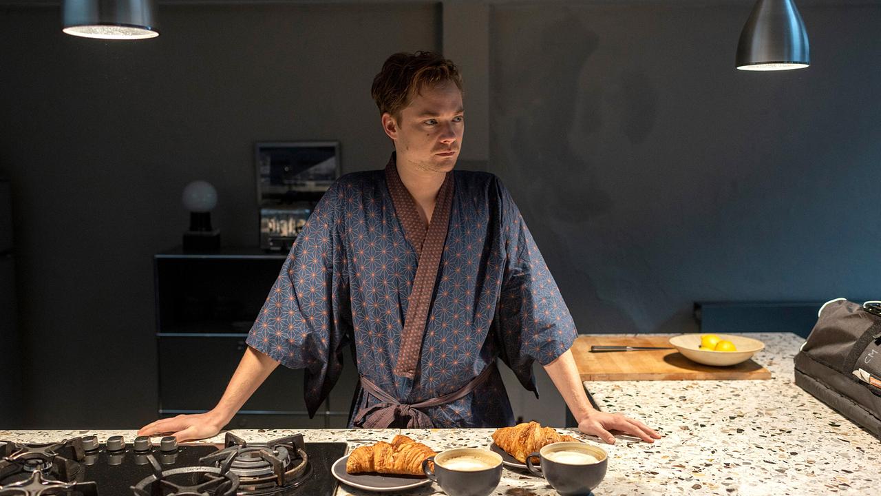 Jan Kuper im gemusterten Morgenmantel steht in einer modernen Küche vor Kaffee und Croissants.